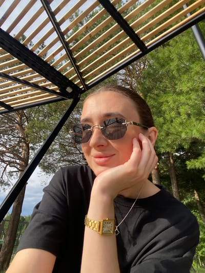 A woman is photographed outdoors, likely at a park or recreational area, near the city of Surgut, Russia. She is leaning on her hand, wearing a black t-shirt, sunglasses, and a gold watch. The background shows trees and a wooden canopy structure. The lighting suggests it is daytime, with sunlight filtering through the trees.