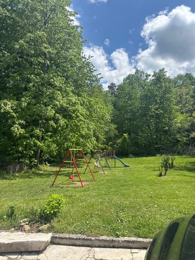 The image presents a residential backyard located in Barziya, Bulgaria. The yard is covered in green grass. A playset with a slide and swings is located near the center of the frame. Trees border the yard, with the sky visible above the tree line. A car is partially visible on the right edge of the frame, near a concrete structure.