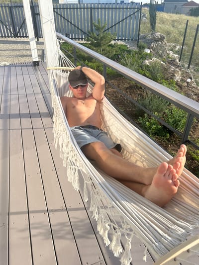 A young man is lying in a hammock outdoors. He is wearing a black baseball cap, sunglasses, and grey shorts. The hammock is made of white rope with tassels. He is on a wooden deck, with a metal railing behind him. In the background, there is a fence, some rocks, greenery, and a building. The sun is shining, indicating it is daytime, and the weather appears to be warm and pleasant, suitable for relaxing in a hammock. The overall mood is one of relaxation and leisure. There is no visible text in the image. The location context provided is Мысовское сельское поселение, Ukraine.