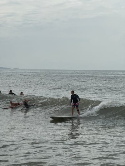 In Phan Thiết, Vietnam, several surfers are in the ocean on a gray, overcast day. One surfer, wearing a dark long-sleeved rash guard and patterned shorts, is actively riding a wave on a surfboard. In the background, other surfers are either paddling on their boards or partially submerged in the water. The sea is choppy, with visible waves and whitewater. The horizon line is flat, with a hazy sky above the ocean. A distant shoreline or landmass is visible on the left side of the image.