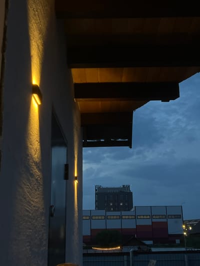 The image depicts a nighttime scene in an urban or semi-urban setting, possibly a courtyard or rooftop area. The foreground features a textured surface, likely concrete, illuminated by several small, bright lights. These lights cast a warm glow on the ground, highlighting the rough texture.
In the mid-ground, there is a dark, rectangular opening or pool of water, reflecting the dim ambient light and the sky above. Two more of the small, bright lights are visible on the surface next to this opening, one casting a strong beam upwards and to the left.
The background reveals structures that suggest a built environment. To the left, a dark, vertical structure with horizontal wooden planks is partially visible, with some light reflecting off its surface. Further back, several multi-story buildings are present. One prominent building consists of stacked, rectangular modules with windows, alternating between white and red-brown sections. Another taller, darker building with visible windows is to the left of this modular structure. The sky above is a deep blue with wispy clouds, indicating nighttime. String lights are visible along the edge of one of the background buildings on the right.
The overall mood is quiet and perhaps a bit mysterious due to the low light and the darkness. The presence of artificial lighting suggests an inhabited or maintained space, despite the lack of visible people. The time of day is clearly night, and the weather appears to be clear, allowing for visibility of the sky and distant structures. The location context provided is Солонцы, Russia. No text is visible in the image.