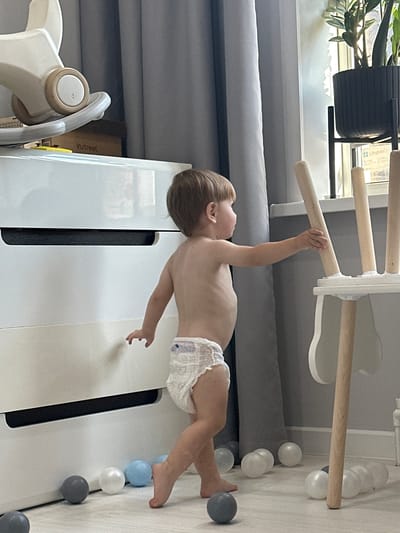 The image shows a toddler with brown hair, wearing a diaper, hanging from a wooden bar of a wooden indoor climbing structure. The structure has white frames and multiple wooden bars. The toddler's body is positioned horizontally, with their right arm extended upwards and grasping the bar. Their left arm is bent, and their legs are slightly bent. The toddler appears to be looking to the left, with their head turned.
The setting is indoors, likely a playroom or nursery. The floor is a light-colored wood or laminate. Scattered on the floor around the climbing structure are several small plastic balls in white, gray, and light blue. In the background, there are gray curtains and what appears to be a white piece of furniture. To the left of the climbing structure, there is a black pot with a green plant.
The lighting suggests it is daytime, possibly morning or afternoon, with natural light coming from an unseen window. There are no strong indications of the weather or specific emotions. The image is taken from a slightly low angle, looking upwards at the toddler.
The text "in:street" is visible on a yellow label on a cardboard box in the background. The location context provided is Эстачи, Russia.
