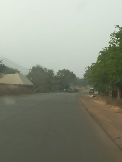 The image displays an asphalt road scene in Kerang, Nigeria, under a hazy, overcast sky. The dark grey road, lacking visible markings, extends into the distance, curving moderately to the right in the midground. On the left side of the road, a building with tan walls and a light-colored, pyramidal roof is positioned. Beyond this structure, dense green and brown foliage, alongside a utility pole with wires, obscures the midground, with faint, distant mountains visible against the sky. A single individual on a dark two-wheeled vehicle, consistent with a motorcycle or bicycle, is observed traveling away from the viewer on the road in the mid-distance. The right side of the road features a dirt shoulder. A cluster of vehicles, including multiple light-colored cars or vans and a white van or bus, are visible stationary on the right shoulder and adjacent to the road curve. Large green trees line the right side of the frame, extending upwards. Indistinct objects or signage are present on the ground near the white van.