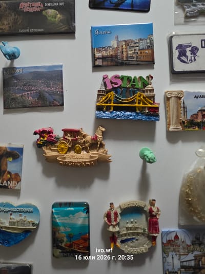 The image displays a collection of refrigerator magnets on a white magnetic surface. Several magnets feature city names and landmarks. From top left, a magnet shows "DEVETASHKA CAVE - ПЕЩЕРА" with Cyrillic text below reading "БЪЛГАРИЯ - BULGARIA", suggesting a location in Bulgaria. To its right, a magnet depicts "Girona". Below that, a magnet with "ISTANBUL" in colorful letters is visible. Further down, a magnet for "Heidelberg" is present, and another magnet features a horse-drawn carriage. Another magnet displays a cow and the text "LAND". Towards the bottom, a heart-shaped magnet reads "OPJE-MACEDONIA". Next to it, a magnet depicts a coastal scene with buildings and palm trees, and text partially visible as "ANTALYA". To the right of that, a magnet has the name "Ivo.wf" and the date "16 юли 2026 r. 20:35". Other magnets show architectural elements and figures. The overall environment is a plain, white, vertical surface, likely a refrigerator door. The lighting suggests it is an indoor setting.
