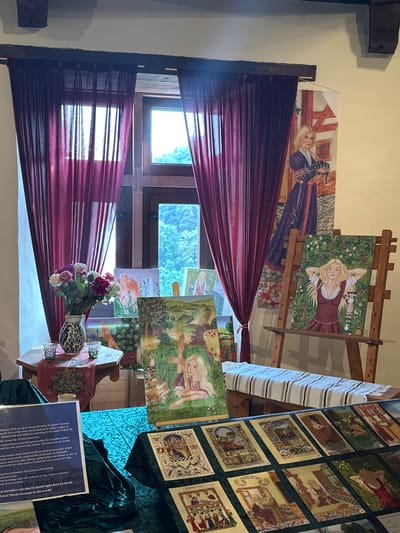 This image depicts an indoor exhibition space, likely a room within the Vianden Castle in Vianden, Luxembourg, given the style of the interior and the presence of historical-themed artwork. The scene is set up as a display for paintings and art prints. In the foreground, a dark green velvet cloth covers a table displaying a grid of rectangular art prints with detailed illustrations, possibly tarot cards or historical scenes. To the left, a rectangular blue plaque with white text provides information about an artist named Norman. Behind this, several paintings are arranged, including a vibrant landscape with poppies and a painting of a woman with blonde hair lying in a field of flowers, displayed on a wooden easel. Further back, a vase of artificial flowers sits on a hexagonal wooden table next to two small glasses. Large, sheer burgundy curtains hang from a dark wooden beam, partially obscuring two windows that offer a view of lush green foliage outside. To the right of the curtains, a large banner-style artwork hangs on the wall, featuring a woman with blonde hair holding a striped cat, dressed in a long purple robe. The overall ambiance is that of an art exhibition with a focus on historical or fantastical themes.