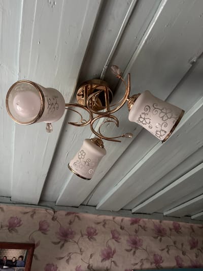 A close-up, low-angle shot captures a decorative ceiling light fixture with three glass lampshades, each adorned with a floral pattern and accented with gold trim. The fixture is attached to a light blue, slatted wooden ceiling. Below the ceiling, a floral wallpaper with pink and purple blossoms lines the wall. In the bottom left corner, a framed photograph shows a group of people. The scene appears to be indoors, likely in a residential setting in Відзаўскі сельскі Савет, Belarus.
