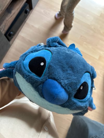 A close-up, eye-level shot shows a fuzzy blue plush toy, identified as Stitch from Disney's Lilo & Stitch, being held by a person wearing a tan shirt. The toy is positioned in the foreground, filling most of the frame with its large, black eyes and prominent blue nose. In the blurred background, a child wearing light brown pants and pink slippers walks across a light brown wooden floor. To the left, a dark brown wooden entertainment center with black hardware is partially visible. The scene appears to be indoors, likely in a residential setting within the city of Waldbröl, Germany.