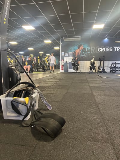 The image depicts the interior of a fitness center or gym, located in Wolfisheim, France. The floor consists of black interlocking rubber mats. The ceiling is a black tiled false ceiling with multiple square-shaped fluorescent light panels providing illumination.
In the immediate foreground, to the left, a clear plastic storage bin is positioned on the floor, containing a black weightlifting belt with a yellow handle, a separate black strap, and other unidentified gym accessories. A black weightlifting belt is partially spilled out from the bin onto the floor. To the far left, a portion of a black and yellow Concept2 rowing machine is visible.
In the mid-ground, a person with light brown hair, wearing a black t-shirt and blue shorts, is seated and operating a chest press machine, which is predominantly yellow and black. Further to the right, a male individual in a white t-shirt and light shorts stands, holding a white towel or cloth in his right hand. To the far right, another person in dark clothing is seated on a black Concept2 rowing machine, facing away from the viewer. A second, identical Concept2 rowing machine stands unoccupied next to it.
The background wall features a large mural depicting a woman exercising, with text partially visible that reads "CROSS TRAININ". Below the mural, the wall has a metallic, corrugated paneling texture. A red fire extinguisher is mounted on the wall to the left of the metallic paneling. Various other yellow and black weight machines are visible further in the background, lining the perimeter of the gym space.