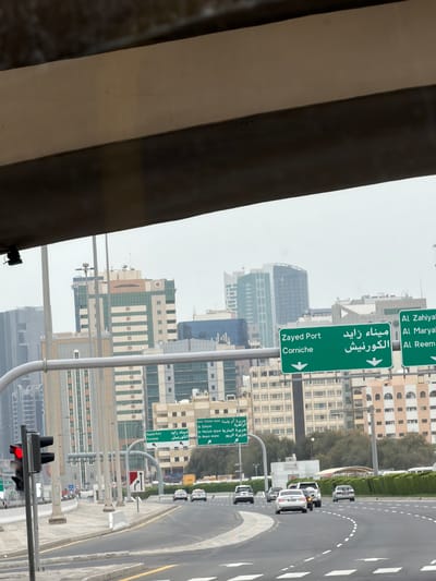 The image captures a street view in Abu Dhabi, United Arab Emirates, likely taken from within a vehicle. Dominating the scene are green directional road signs in both Arabic and English, indicating directions to Zayed Port and Corniche, as well as Al Zahiyah, Al Maryah, and Al Reem. Several high-rise buildings can be seen in the background. The road is visible, with cars driving, and there is a traffic light in the foreground with a red light.