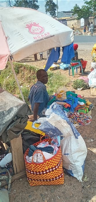 This image captures an outdoor market scene in Kipkarren, Kenya, under bright daylight. The ground is unpaved and dusty, suggesting a roadside or informal market setup.

The central figure is a man, likely a vendor, wearing a blue short-sleeved shirt. He is standing or leaning amidst his merchandise, looking towards the viewer's right with a neutral expression.

To the left, a large, light-colored canopy or umbrella tent partially shades the area. Prominently displayed on the canopy is the word "NGAAE" in red, along with "TUIHUANE MASHINANI" and a logo depicting intertwined figures, indicative of a community or organizational presence. Beneath the canopy, several blue garments are hanging, possibly trousers or uniforms offered for sale.

The man's stall is laden with various goods. To his immediate right, a large white and blue patterned sack is on the ground. Next to it, a striking woven basket with an orange, yellow, white, and black checkerboard pattern is filled to the brim with textiles or clothing. Behind him and to his left, there are piles of colorful fabrics (including blues, greens, and reds) and a assortment of other small items, some appearing to be packaged goods or plastic articles. A green plastic chair is also visible in the background.

The strong sunlight creates visible shadows, indicating it is likely mid-morning to afternoon. The scene depicts the everyday activity of a local street vendor selling a variety of items in an informal market environment.