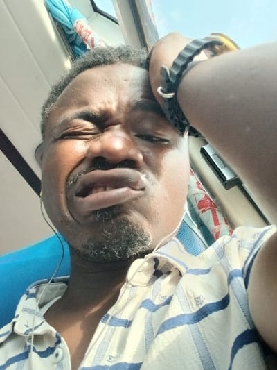 The image shows a close-up of an African man's face. He appears to be in a state of distress, with his hand on his forehead and his mouth slightly open. He is wearing a striped white and blue shirt, a black wristwatch, and headphones. He is sitting in a vehicle. The vehicle has a blue seat and a window. The location is a public transport vehicle in New Karu, Nigeria. The coordinates are 8.954 latitude and 7.679 longitude.