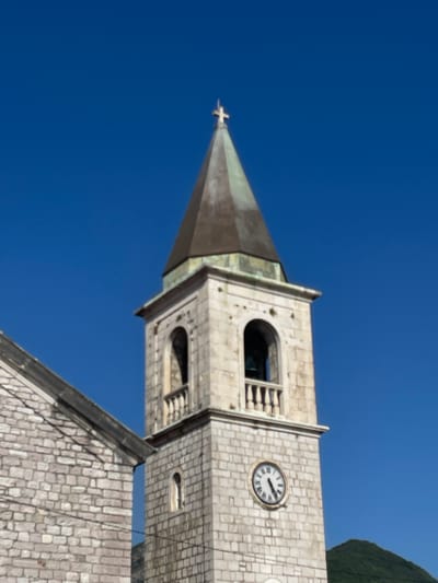 This is a low-angle, upward shot of a church tower against a clear blue sky. The tower is made of light-colored stone blocks and has a conical, dark-colored spire topped with a cross. A clock face with Roman numerals is visible on one side of the tower. The clock hands indicate approximately 3:55. The scene suggests a sunny day. The stone architecture, including the cross and the clock, are typical elements of a church. The overall impression is one of architectural grandeur against a serene sky. The image seems to be taken in Donja Lastva, Montenegro, as indicated by the context provided. There is no visible text in the image itself.