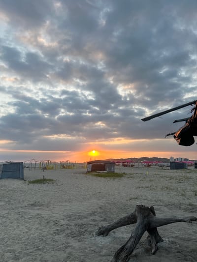 The image captures a sunset over a beach in General Villamil Playas, Ecuador. The sky is filled with gray clouds illuminated by the setting sun, which appears as a bright orange orb on the horizon. The beach is sandy and slopes gently. Various beach structures are visible, including sunshades. In the upper right corner, a weathered piece of driftwood rests on the sand. A fragment of an unidentified object hangs in the top left corner.