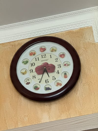 A wall clock with a dark brown frame is mounted on a textured, pale yellow wall. The clock face is white and features colorful illustrations of fruits at each hour mark, along with Arabic numerals. The hands of the clock are black, with the hour hand pointing approximately to 9 and the minute hand pointing approximately to 7. The brand name "GOTIME" is visible on the clock face. To the left of the clock, a decorative white molding runs vertically along the edge of the wall. The overall scene suggests an indoor setting, likely a home in Chemerne, Ukraine. There are no people or specific activities depicted in the image.