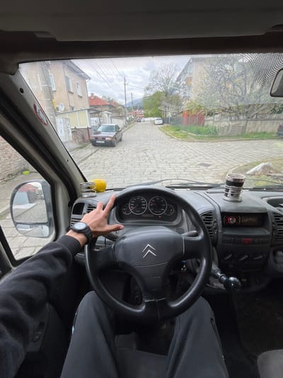 A person is driving a Citroen vehicle on a cobblestone street in Sopot, Bulgaria. The person wears a black watch and a dark sweatshirt. A yellow lemon sits on a holder on the dashboard. A dark-colored Audi vehicle is parked on the side of the street. Buildings are visible outside the windshield. The sky is overcast.