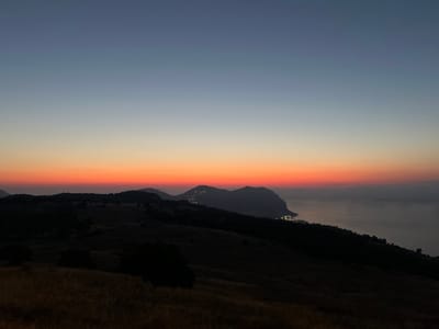 A wide-angle, landscape photograph captures a dramatic sunset over the Mediterranean Sea, with silhouettes of mountains and a coastal town in the distance. The sky transitions from a deep twilight blue at the top to a vibrant band of orange and red just above the horizon. The foreground is dominated by dark, undulating hills covered in dry grass. In the mid-ground, the dark, jagged silhouettes of mountains rise from the land. Along the coast, nestled within the mountains, a town is illuminated by scattered lights, hinting at a settlement. The sea itself is a dark, calm expanse meeting the hazy horizon. The overall scene evokes a sense of quiet grandeur and the end of the day. This view is likely from a vantage point in the hills surrounding Palermo, Italy, looking out towards the sea.