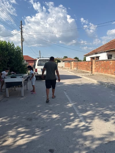 The image depicts a street scene in Vinitsa, Bulgaria, under a bright, partly cloudy sky. A man, viewed from behind, walks down the asphalt road. To his left, two young girls are engaged in a game of foosball on a table placed on the side of the road. A white van is parked behind the foosball table. The street is lined with houses constructed with brick walls and red tile roofs, with a few trees and bushes visible. Electrical poles with numerous wires are prominent along the roadside. The overall scene suggests a typical day in a residential area.