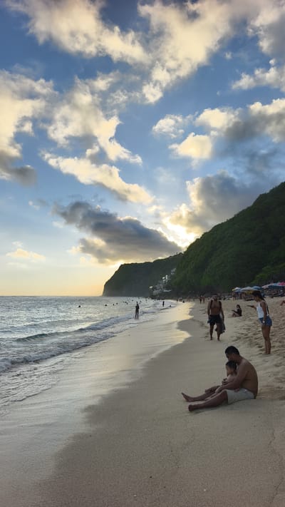 The image depicts a beach scene in Ungasan, Indonesia. The beach is sandy, with gentle waves lapping at the shore. Several people are scattered along the beach, including some swimming in the ocean, walking along the shoreline, or relaxing on the sand. In the foreground, a man sits on the sand with a young child in his lap. A woman in denim shorts and a white top is to the right of them. A man in black shorts is walking away from the water. The background features a lush, green hillside extending along the coast. A break in the hillside reveals a series of white buildings that look like small resorts. The sky is partly cloudy, with the sun creating a soft glow behind the clouds, suggesting it's either sunrise or sunset. The overall mood is peaceful and serene.
