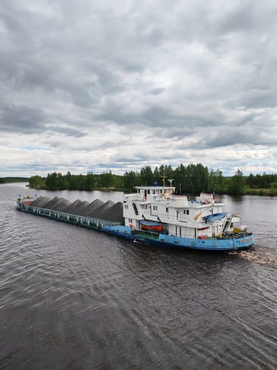 A barge carrying a load of dark gray material, possibly gravel or sand, is moving down a wide, calm river. The barge is long and flat, with a white superstructure at the rear containing multiple decks and windows. The superstructure is painted with blue trim and has life preservers visible on the exterior. The barge appears to be propelled by a tugboat, though only the rear of the tug is visible, creating a wake in the water.
The riverbanks on either side are lined with dense, green trees. The sky above is overcast with a dramatic mix of gray clouds, suggesting impending or recent rain. The overall atmosphere is one of industrial transport occurring in a natural, somewhat remote setting. The location is identified as Ozernoe Ust'e, Russia.