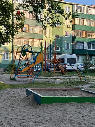A residential building in Zheleznogorsk, Russia, features a playground in the foreground. The building, painted a textured green, has multiple floors with balconies and windows. A metal playground structure with a swing set and a slide dominates the middle ground. Behind the playground, cars are parked, including a white van. A sandbox, partially filled with sand and bordered by blue and green wood, is visible in the lower foreground. The scene is set outdoors, with trees and grass around the playground area.