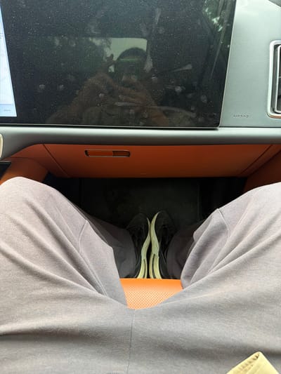 The image shows a first-person perspective of a person sitting in a car. The view is focused on the person's legs and feet, which are clad in grey sweatpants and black and white sneakers. The person's hands are visible reflected in the car's touchscreen, holding a phone. The car's interior features an orange glove compartment and grey trim. The touchscreen displays a map with the word "Шоссе" (Highway) visible, indicating a location in Russia. The "AIRBAG" warning is also visible on the dashboard. The image appears to have been taken in Zhukovka, Russia, due to the Cyrillic text on the screen. The overall scene is mundane and static, with no apparent action or interaction taking place beyond the reflection of the person taking the photo.