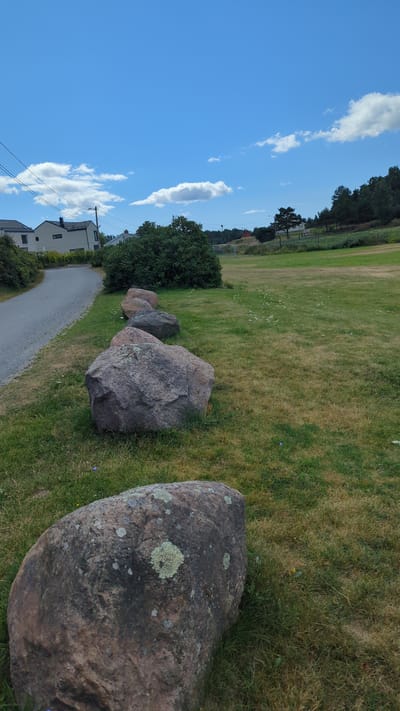The image shows a clear blue sky with scattered white clouds over a green grassy field in Larkollen, Norway. A paved path runs along the left side of the image, bordered by a row of large, lichen-covered boulders. Beyond the path, there are houses and trees. The field extends into the distance, with a treeline visible on the horizon. The overall scene is serene and natural, with a hint of suburban development.