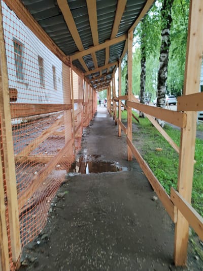 The image shows a temporary walkway covered by a wooden and metal structure, likely a construction shed or scaffolding, set up along the side of a white building. The walkway is paved with asphalt and has puddles of water, suggesting recent rain. The building has multiple windows and is partially obscured by orange safety netting attached to wooden posts, indicating ongoing construction or renovation.
To the right of the walkway, a wooden fence with horizontal planks separates the path from a grassy area with several birch trees. In the background, more trees and some parked cars are visible, suggesting an outdoor, urban or suburban environment. A single person is walking away from the camera down the covered walkway, their figure small in the distance, adding a sense of scale to the scene.
The overall mood is subdued, likely due to the overcast or rainy weather indicated by the wet ground and puddles. The time of day appears to be daytime, as there is sufficient natural light. The location context points to Syktyvkar, Russia. No visible text is present in the image. The scene captures a moment of passage under a temporary shelter, perhaps for pedestrian safety or protection from the elements during construction.