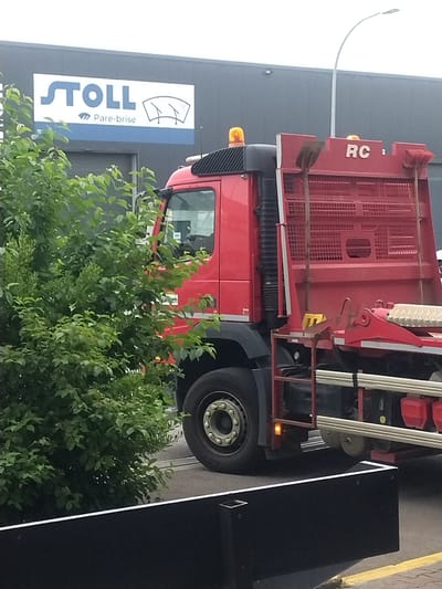 The image, taken in daylight, features a red specialized truck parked in what appears to be a commercial or industrial area in Leudelange, Luxembourg. The truck has a bright red cab and a robust red body equipped with metal grating and various tools, including a shovel attached to the rear. The white letters "RC" are visible on the truck's rear body. Yellow warning lights are present on the cab and side of the vehicle.
In the foreground, a dense green bush partially obscures the lower front part of the truck. Behind the truck, to the left, is a light grey building with a large sign. The sign reads "STOLL" in prominent blue letters, with a graphic of windshield wipers above and the French words "Pare-brise" (meaning "windshield") below, indicating a glass repair or replacement business. A curving street lamp is visible extending from the upper left. To the right of the truck, a tall, black, rectangular structure stands next to a road or parking area, which is delineated by a yellow line on the asphalt. The lighting suggests an overcast or bright cloudy day. No people are visible, and the truck appears stationary.