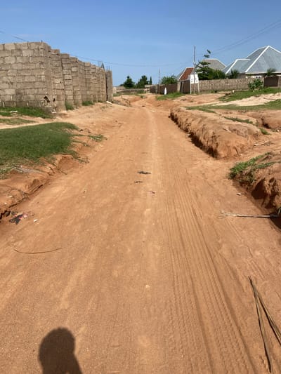 The image depicts a sun-drenched scene in Kuson, Nigeria, likely in the late morning or early afternoon given the strong shadows. The central focus is a dirt road, its surface marked with distinct tire tracks, suggesting recent vehicle activity. The road is flanked by areas of uneven, dry earth, with patches of green grass growing along the edges and in some of the eroded sections.

To the left of the road, a partially constructed building is visible. It appears to be made of grey cinder blocks, stacked in an unfinished state, with some exposed rebar at the top. Beyond this construction site, several houses with distinctive pitched roofs, some metallic and others appearing to be tiled, are nestled amongst greenery. Electrical wires stretch across the scene, supported by wooden poles, indicating the presence of infrastructure.

The overall setting is rural or semi-urban, characterized by the natural dirt road, the ongoing construction, and the residential buildings. The weather appears to be clear and sunny, as evidenced by the bright sky and the sharp shadows cast by objects and the photographer. There is no visible text within the image. The scene conveys a sense of development and daily life in a Nigerian community.