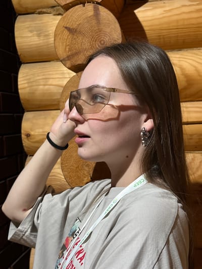 A young woman with long brown hair, wearing oversized sunglasses, a beige t-shirt, and a silver earring, poses in front of a wooden wall. She has a black hair tie on her wrist. A lanyard with the logo "SBER" is visible around her neck. The background is a wall constructed of large, round wooden logs. The lighting suggests an indoor environment, possibly a rustic building or a sauna, given the wooden construction. The image is taken from the chest up, with the woman looking to her left. The presence of the "SBER" logo indicates the location is likely within Russia, possibly related to a Sberbank event or location. While Elovo is a rural settlement in Perm Krai, Russia, this image provides no specific visual cues to confirm that exact location beyond the general context of Russia suggested by the Sberbank branding.
