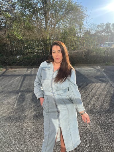 A single female subject stands on a dark asphalt surface in Montigny-le-Bretonneux, France. She has long, dark brown hair and a neutral facial expression. The subject is wearing a light blue, long-sleeved denim jumpsuit with a full-length front zipper, chest pockets, and side pockets; her left hand is inserted into a pocket. The background features a metal fence partially obscured by dense deciduous trees and shrubs showing early spring foliage. A light-colored vehicle and a dark cylindrical bollard are faintly visible in the far background beyond the vegetation. Strong, direct sunlight from the upper right of the frame casts defined shadows on the paved ground.