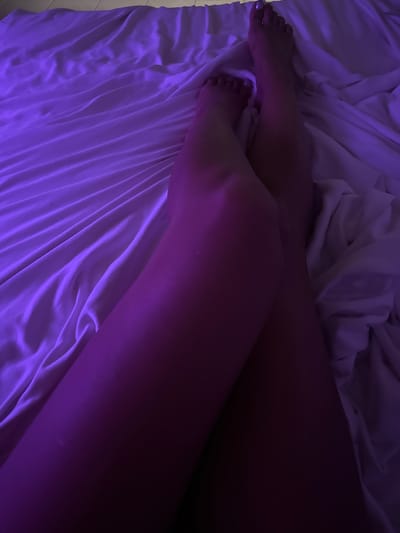 The image shows a close-up, overhead view of a person's legs crossed and lying on white sheets. The lighting is predominantly purple, casting a shadow across the lower portion of the legs. The sheets are rumpled, suggesting they are in a bed. The setting is domestic and appears to be indoors. There are no visible elements that indicate a specific location in Peleana, Sri Lanka.
