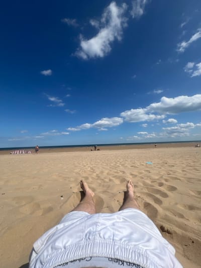 A sunny beach scene shows a blue sky with scattered white clouds. The sand is light brown, marked with footprints. A pair of feet are visible in the sand, and white Calvin Klein swim trunks dominate the lower right. The ocean appears as a thin line in the distance, and a few people can be seen on the beach. The image is rotated ninety degrees.