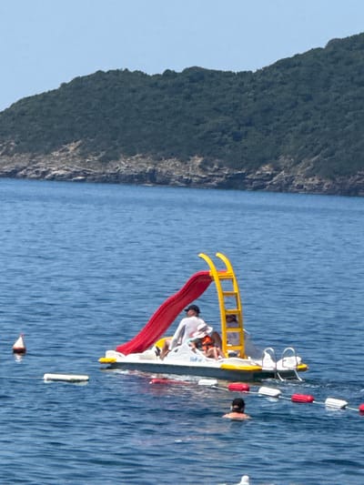 In Kamenovo, Montenegro, a pedal boat with a red slide and yellow ladder is situated on the calm blue sea. Two individuals are visible on the boat: one person, wearing a white shirt, is seated near the slide, and another, wearing an orange life vest and a straw hat, is also seated. A third person is swimming in the water nearby, with buoys marking a boundary in the sea. In the background, a lush green, tree-covered mountain slopes down towards the rocky coastline, with a clear blue sky overhead.