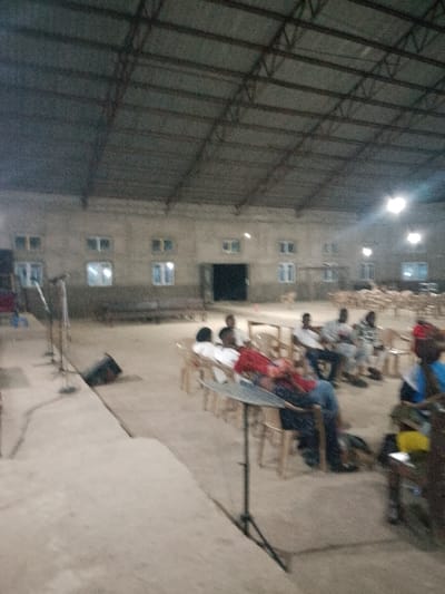 The image depicts an indoor gathering in a large, unfinished building, possibly a community hall or a place of worship, located in Serti, Nigeria. The structure has a corrugated metal roof supported by exposed beams and has plain concrete walls with rows of windows. The floor is also concrete.
In the foreground, there is a raised platform or stage area with microphones and sound equipment set up. Several individuals are seated in chairs facing the platform. The people appear to be attending an event, possibly a sermon or a presentation, as suggested by the setup and their seated posture.
The lighting in the building seems to be artificial, with some light fixtures visible on the ceiling. The overall atmosphere is one of a functional but not elaborately decorated space, typical of community-oriented venues in the region. The people present are casually dressed, and their interactions are not clearly discernible due to the distance and image quality. The scene suggests a communal gathering for a specific purpose within a local context.