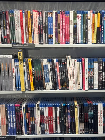 The image captures three tall, densely packed vertical stacks of Blu-ray and DVD cases, likely displayed on shelves in a store or a personal collection. The media cases are predominantly blue-spined Blu-rays, with many still encased in their original plastic wrap, indicating new or well-preserved items.
The titles visible on the spines include a mix of French and English language films. In the leftmost stack, titles such as "MOONLIGHT EXPRESS," "MONSIEUR HIRE," "MISERY," "LE MÉLANGE DES GENRES," "MEAN STREETS," "MA VIE MA GUEULE," "Materialists," "Ma Mère, Dieu et Sylvie Vartan," "MADRES PARALELAS," "MAD DOG MORGAN," "LOVE LIES BLEEDING," and "LOST HIGHWAY" are legible.
The middle stack features "RAMBO III," "RAISON & SENTIMENTS," "RAGTIME," "LA PERSONNE," "Quand Harry rencontre Sally...," "EL PROFESOR," "LE PRIX DU DANGER," "PILE OU FACE," "PHOENICIAN," "PATRIOTES," "PÉPÉ LE MOKO," "PARIS, TEXAS," and several "PAGNOL FERNANDEL" titles.
The rightmost stack displays "Les Superflics de Miami," "SUBWAY," "SUBSERVIENCE," "THE STRANGER," "SPENCER," "SORRY," "SONGS OF LOVE," "SOLDAT COLLINS," "SOIS-BELLE et tais-toi !," "SISTER ACT," "SIBERIA," "SHOCK CORRIDOR," "SHOWING UP," "SHE SAID," "SHADOW FORCE," "SENS UNIQUE," "SECRET CHAMBRE NOIRE," and "LES SCHTROUMPFS LE FILM." Several titles, like "SISTER ACT" and "LES SCHTROUMPFS LE FILM," appear to have multiple copies.
The scene is brightly lit, suggesting an indoor environment. No people are visible, and the focus is entirely on the extensive collection of films. The background is a plain, light grey surface. The arrangement implies an organized display of cinematic works.