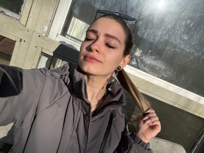 A close-up selfie depicts a young adult female outdoors in Reutov, Russia. She has light brown hair pulled back into a high ponytail, with black-framed sunglasses resting on top of her head. Her eyes are closed, and her face is directed upward, seemingly towards the sun, which casts strong illumination. She is wearing a grey, zippered puffer jacket. Her left hand is positioned near her left shoulder, gently grasping a section of her ponytail, and her fingernails display a light, glittery polish. A dark ear gauge or plug with an attached dangling earring, featuring small, light-colored beads, is visible in her left ear. The background consists of a weathered building facade featuring multi-pane windows. The window frames are constructed from light-colored wood with noticeable peeling paint. The glass panes reflect bright sunlight and the indistinct, bare branches of trees.