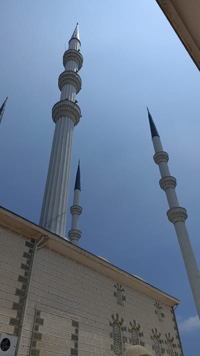 This is a low-angle, eye-level shot of the minarets of a mosque in Yenişehir, Turkey, against a clear, pale blue sky. The foreground features the textured, light-colored brickwork of the mosque's main building, with decorative grey accents and arched windows featuring intricate lattice work. To the right, a tall, slender white minaret with multiple decorative bands rises vertically. In the middle ground, another, slightly shorter white minaret with similar decorative bands stands. Further in the distance, two dark, conical minarets with decorative bands pierce the sky. A thin wire stretches diagonally across the frame between two of the minarets. The scene suggests a bright, sunny day.
