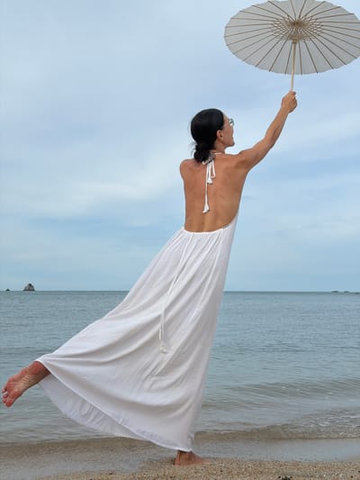 A woman stands on a sandy beach, her body angled away from the camera, and her left arm is raised, holding a white parasol against a cloudy sky. She wears a flowing white dress with a low back and thin straps. Her feet are bare, with the left foot lifted and visible beneath the hem of her dress. The ocean is to her right, with small waves lapping at the shore. In the distance, a small rocky island is visible. The scene is set in Baan Plai Laem, Thailand.