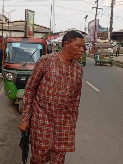 This image captures a daytime street scene in Aba, Abia, Nigeria. A dark-skinned male, appearing middle-aged to elderly, with a short, dark haircut, stands on an asphalt road. He is dressed in a two-piece traditional attire consisting of a long-sleeved shirt and trousers, both patterned with a reddish-brown and beige plaid design. His right hand grips a crumpled black plastic bag, while his left hand is partially obscured, possibly in a pocket. His mouth is slightly ajar, and his gaze is directed towards the right of the frame. To his immediate left, a parked green and dark red/brown Bajaj RE 4S tricycle ('keke napep') is visible. Further down the multi-lane road, two additional green tricycles are present, one stationary and one in motion. The background features a row of urban buildings with corrugated roofs, utility poles, and multiple billboards. One prominent billboard on the right side of the street displays "ABA RIVER MILL LIMITED," confirming the location. A partially visible green billboard is behind the initial tricycle, and a distant banner shows multiple human faces. The road surface is gray asphalt with a faded white lane marker. The sky is light gray, indicating an overcast day.