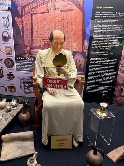 This media file captures a museum display featuring a lifelike mannequin of King Midas seated in a wooden chair, holding a golden mirror. The mannequin is dressed in a light-colored robe with intricate gold and green trim and has a distinct receding hairline and aged facial features.
The scene is set indoors within a museum, characterized by display cases and informational panels. A red sign held by the mannequin prominently warns "DİKKAT! CAM VİTRİN" (CAUTION! GLASS DISPLAY CASE), indicating a glass barrier in front of the exhibit. A small golden plaque beside the mannequin identifies him as "Kral MİDAS / Frig Kralı / Phrygian King."
Behind and around the mannequin, several informational panels are visible. The left panel is titled "MİDAS EFSANELERİ" (Midas Legends) and details two famous myths: Midas's donkey ears and his golden touch. Key phrases like "MİDAS'IN KULAKLARI EŞEK KULAKLARIDIR" (MIDAS'S EARS ARE DONKEY EARS) are discernible. The right panel is titled "YAZILIKAYA" and discusses the Midas Monument, mentioning its location in Eskişehir, Turkey, and its architectural significance.
Various historical artifacts are displayed in front of and beneath the mannequin's exhibit area, some in sand-filled trays, others in clear stands. These include pottery, metal vessels, and wooden ornaments, with labels such as "Toprak Kap" (Earthen Pot), "Törensel İçki Kabı" (Ceremonial Drinking Vessel), "Ahşap Süsleme" (Wooden Ornament), "Göbekli Tas (Tunç)" (Belly Bowl (Bronze)), and "Bakraç (Tunç)" (Copper Pot (Bronze)). The overall display aims to educate visitors about King Midas, Phrygian culture, and local historical sites in Eskisehir, Turkey.