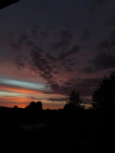 This is a vertical, low-angle, dimly lit photograph of a sunset over an outdoor, rural area. The sky dominates the upper portion of the frame, displaying hues of deep purple, dark gray, and bands of vibrant orange and light blue from the setting sun. Scattered clouds are visible throughout the sky, with some appearing reddish-brown and others dark gray. The lower third of the image consists of a silhouetted landscape. In the foreground, a dark, rectangular structure, possibly a greenhouse or shed with a corrugated roof, is visible. Behind it, the silhouettes of various trees and foliage stretch across the frame, with a more dense grouping of trees on the right side. The background shows a faint outline of distant buildings or structures, suggesting a settlement or village on the horizon. The overall impression is of twilight in a countryside setting.