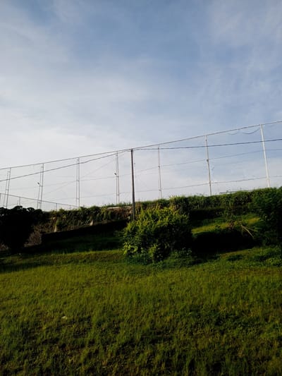 The image depicts a grassy slope in Thalang, Laos, leading to a retaining wall topped with a chain-link fence. Vegetation grows both on the slope and along the top of the wall. A single, dark-colored pole stands along the top of the wall. The sky is blue with some light, wispy clouds. There are no people visible.