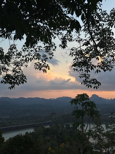 The image captures a landscape in Ban Chiao Lan, Thailand, likely at dusk or dawn given the soft light and colors in the sky. The foreground is dominated by the silhouette of tree branches and leaves, creating a dark contrast against the brighter sky. Through the foliage, the sky displays hues of orange and grey, suggesting the presence of clouds and the transition between day and night.

In the middle ground, a mountainous region stretches across the horizon. The mountain range is dimly lit, blending into the dark environment. Nestled in the valley of the mountains are bodies of water that form part of the Chiao Lan Dam Reservoir. A road or walkway is visible along the reservoir. The general scenery suggests the image was taken from an elevated position, offering a wide perspective of the area. There are no people visible in the image.