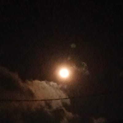 A full moon glows brightly in a dark, cloudy sky, its light illuminating a band of white clouds stretching across the frame. A thin, dark horizontal line, possibly a wire, bisects the image. The overall atmosphere is dimly lit and somewhat obscured by the night and clouds, suggesting a nighttime scene in a location like Cigadung, Indonesia.