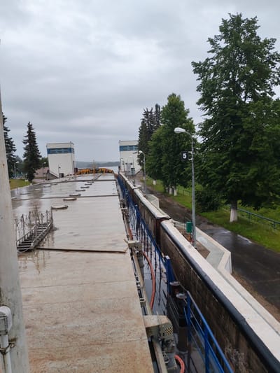 The image depicts the sluice gate of a hydro-engineering facility in Belousovo, Russia. The scene is characterized by an overcast sky and wet surfaces, suggesting recent rainfall. The foreground features a concrete walkway with several metal hatch covers. To the right, a blue metal railing runs alongside the concrete wall of the lock. In the distance, two white buildings with blue-capped windows stand on either side of the waterway. Several mature trees with dark green foliage are visible, some bordering a paved path. A lamppost with a sign displaying the number "5" is positioned near the right edge of the image. The overall atmosphere is industrial and somewhat somber due to the weather conditions.