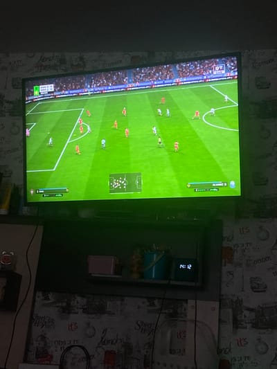 This image shows a television screen displaying a soccer video game, likely FIFA, with a match in progress. The game features players in red and blue uniforms on a green grass field. On the left side of the screen, the crowd is visible, with a "LIVE" indicator and a logo that appears to be for a sports channel or broadcast. Below the gameplay, a scoreline indicates "POR 0" and "ARG 0" with "02:03" underneath, suggesting the current score and time elapsed in the half. On the right side of the screen, a player's name "ALVAREZ 9" is displayed along with their rating and other in-game information.

In the background, the television is placed against a wall with wallpaper that has a repeating pattern of various text and images, including what appears to be London-themed graphics like Big Ben and double-decker buses. A digital clock on a shelf to the right of the TV displays "14:12", indicating the time. The room appears to be dimly lit, with the glow of the TV being the primary light source. The overall scene suggests a casual setting, likely a living room, where someone is playing or watching a soccer game. The image is taken from a slightly angled perspective, and some of the background details are obscured by shadow.