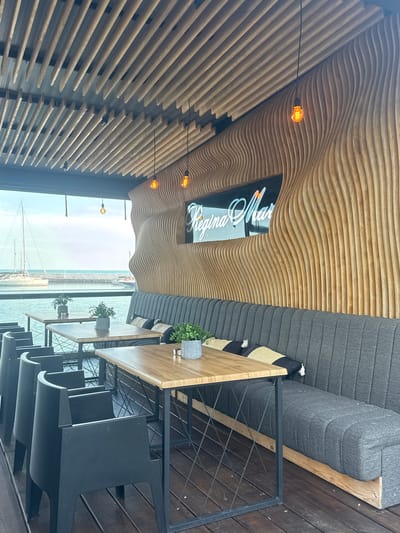 The image shows the interior of a modern restaurant with a view of the sea in Balchik, Bulgaria. The restaurant features a unique interior design with a curved wooden wall and a slatted wooden ceiling. There are several wooden tables with black chairs and a long gray upholstered bench seating area. Potted plants are placed on the tables as decoration. In the background, through a large window, a view of sailboats and the sea can be seen. The restaurant's name, "Regina Mar," is displayed in illuminated lettering on the wooden wall. The overall atmosphere appears to be one of contemporary elegance and waterfront dining.