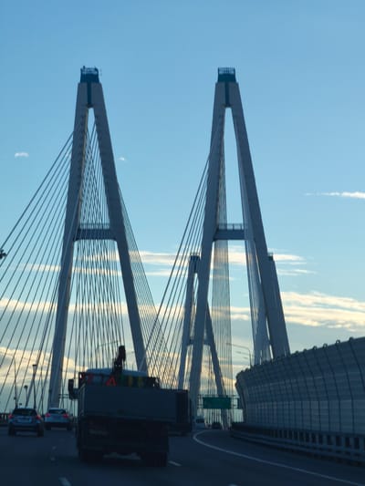 The image shows a view from a moving vehicle on the Big Obukhovsky Bridge, a cable-stayed bridge spanning the Neva River in St. Petersburg, Russia, near the settlement of Novosaratovka. The perspective is from the roadway, looking towards two prominent bridge towers that dominate the frame. The sky is a clear blue with some wispy clouds. The bridge's suspension cables are visible, fanning out from the towers. In the foreground, the rear of a dark-colored truck is prominent, its load obscured. To the left, other vehicles, including cars, are also on the roadway. On the right side of the image, a sound barrier runs along the edge of the bridge. The lighting suggests it is either early morning or late evening.