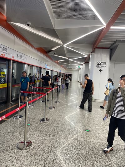 This media file depicts a subway station platform in Panyu District, China. The scene is illuminated by modern, geometric lighting integrated into the ceiling and walls, creating a bright and contemporary atmosphere. The floor is a speckled grey material, and silver stanchions with red velvet ropes mark a waiting line.
Several people are visible, appearing to wait for a train. Some are looking at their phones, while others stand or sit patiently. One man in a black tank top is standing and looking at his phone, and another man in a blue t-shirt is standing near the platform edge. The overall mood seems to be one of routine travel.
Prominent text in the image indicates that this is the "22号线 Line 22" and the direction is "番禺广场方向 Towards Panyu Square." Other visible text includes "州南站 South Railway Station" and "广州地铁 Guangzhou Metro." These details firmly place the scene within the Guangzhou Metro system. The presence of a train door suggests a train is either present or expected imminently. The lighting and clean design imply it is daytime, and the interior setting makes weather irrelevant.